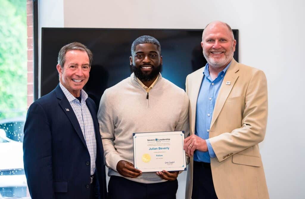 Graduating Fellow holds his certificate between two smiling mentors at a Severn Leadership Group graduation in Annapolis