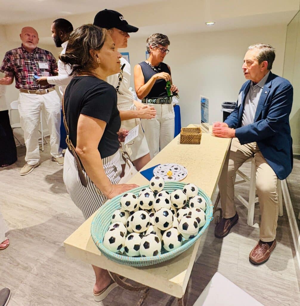 Guest chats with a seated man beside a basket of soccer ball stress balls at a Severn Leadership Group event